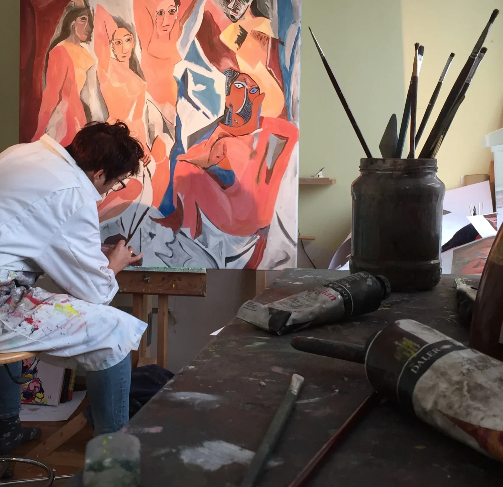 Angelique aan het werk aan een doek in haar atelier, op de voorgrond een pot met kwasten en tubes verf.