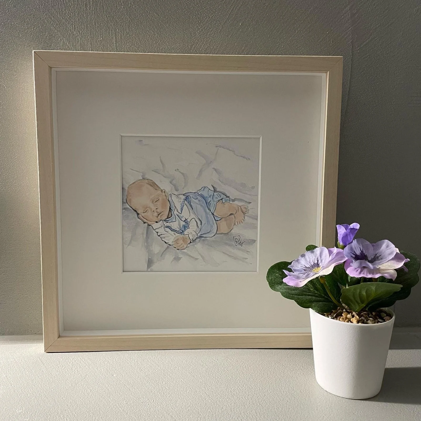Babyportret in aquarel: een liggende baby in een blauw pakje, ingelijst in een blank houten lijst.