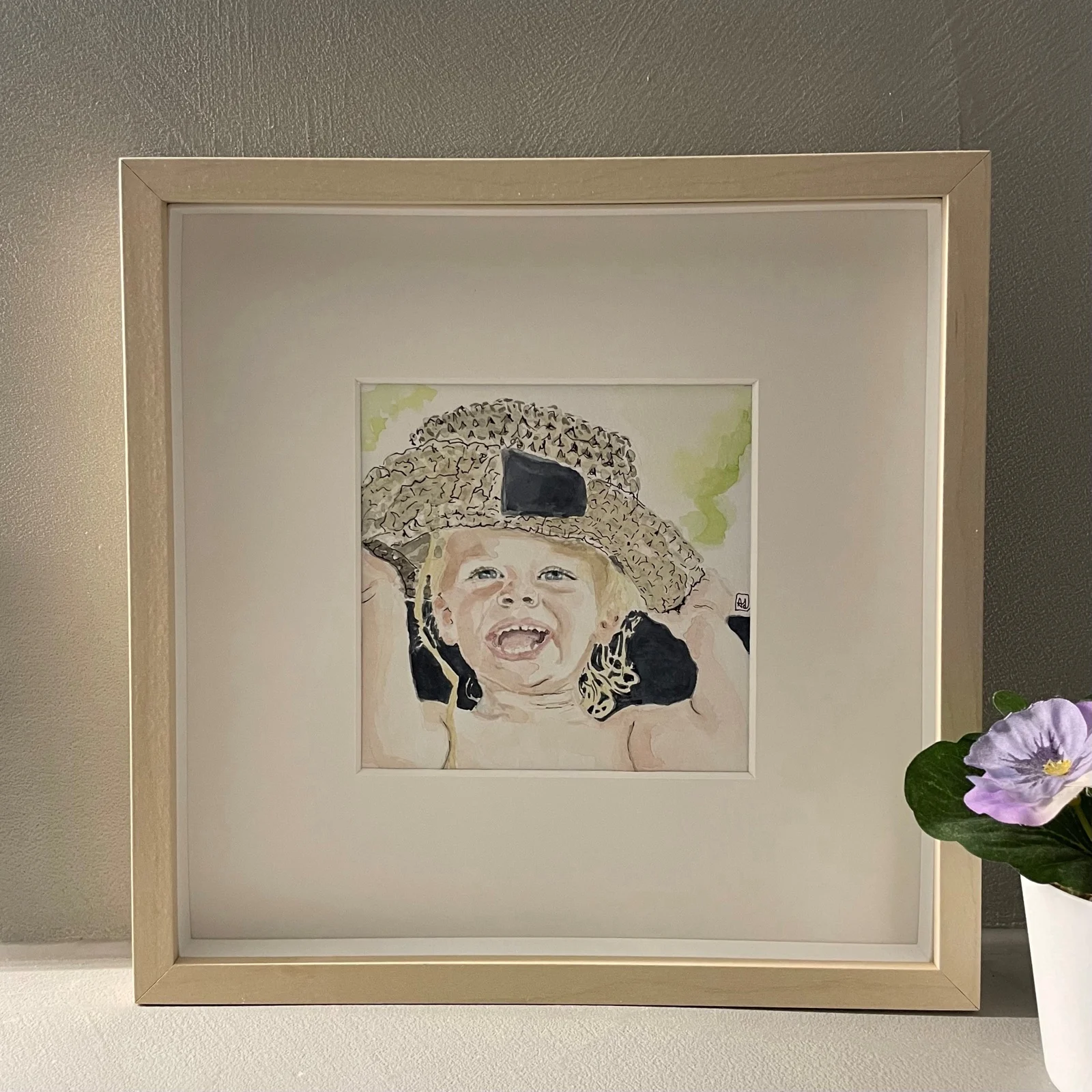 Babyportret in aquarel: een lachend kind met een gehaakt hoedje, ingelijst in een blank houten lijst.