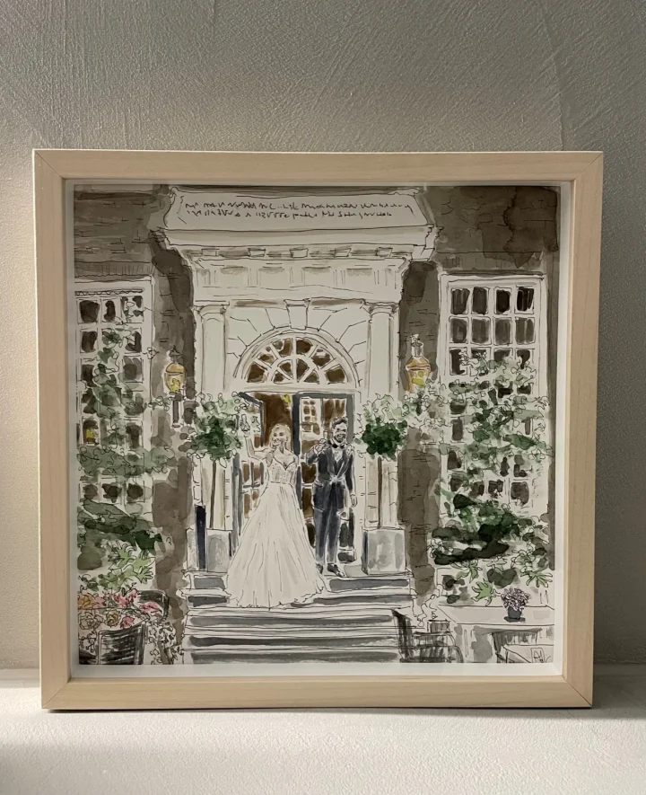 Huwelijksportret in aquarel: een bruidspaar op de trap voor een wit gebouw, ingelijst in een blank houten lijst.