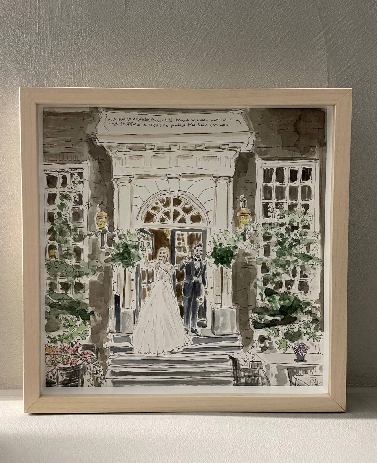 Huwelijksportret in aquarel: een bruidspaar op de trap voor een wit gebouw, ingelijst in een blank houten lijst.