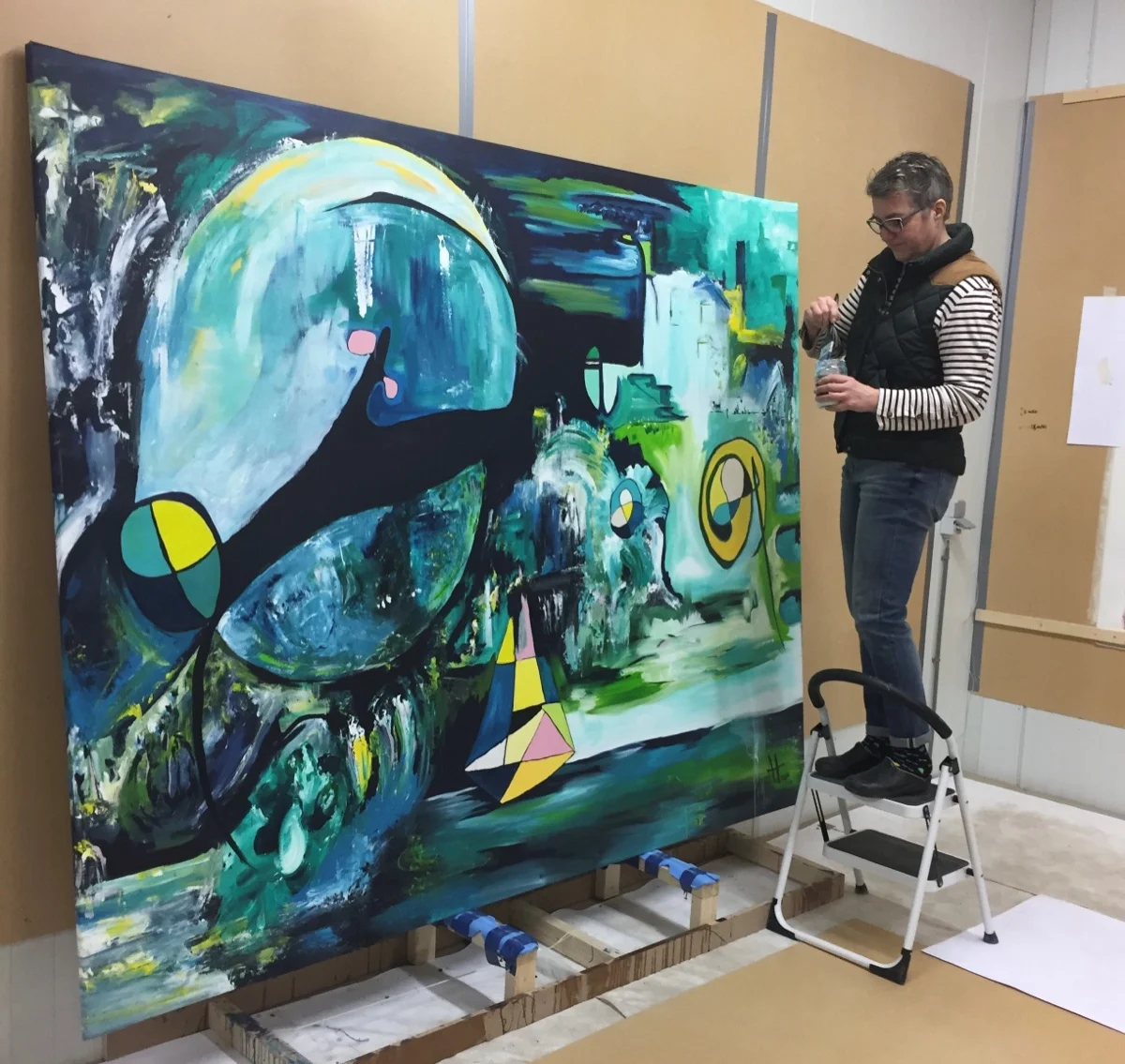 Angelique van den Hogen op een trapje in haar atelier, met een kwast in de hand voor het grote doek Universum.